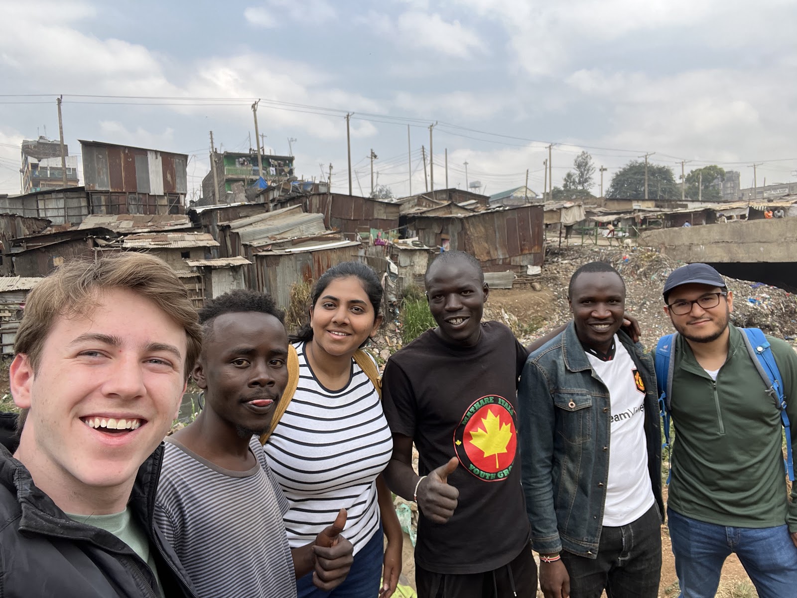 a group of six people posing for a selfie. All are smiling and standing in front of an informal settlement, with makeshift structures and debris visible in the background under a cloudy sky.