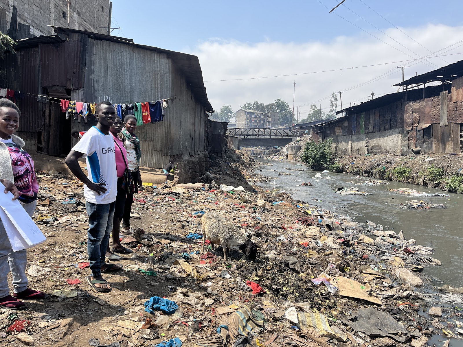 an informal urban area. In the foreground, four individuals are standing alongside a polluted riverbank. All are facing the camera with neutral expressions. The river beside them is littered with waste, and the banks are strewn with trash. Makeshift homes with corrugated metal surfaces and hanging laundry are in the background, and a bridge is visible in the distance. It's a sunny day with a clear blue sky.