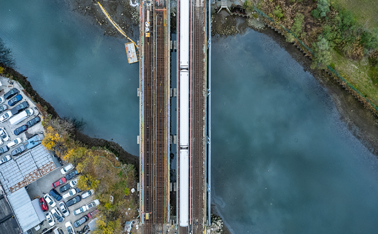 a train bridge over a river. Clouds are reflected in the water. In the upper right corner is the edge of land with green grass and trees. In the bottom left corner is land with some trees and about twenty cars and an industrial building.