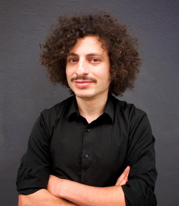 a person in a black collared shirt with mustache and curly hair looking at the camera 