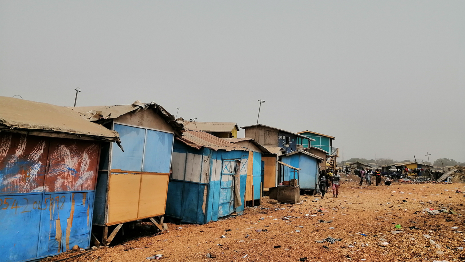 A row of shelters built from colorful sheets of metal with tan rooves extends into the distance. The rocky tan ground has trash scattered across it.